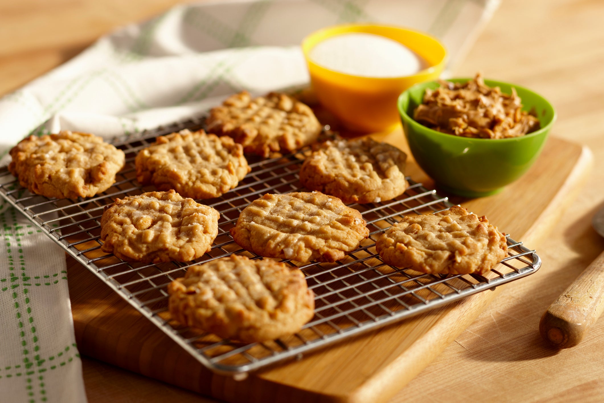 Baked cookies on a cooling rack with a bowl of sugar and a bowl of oats on a wooden surface.